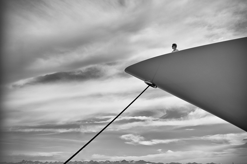 Man standing at bow of yacht, looking out to mountains, open water, and clouds